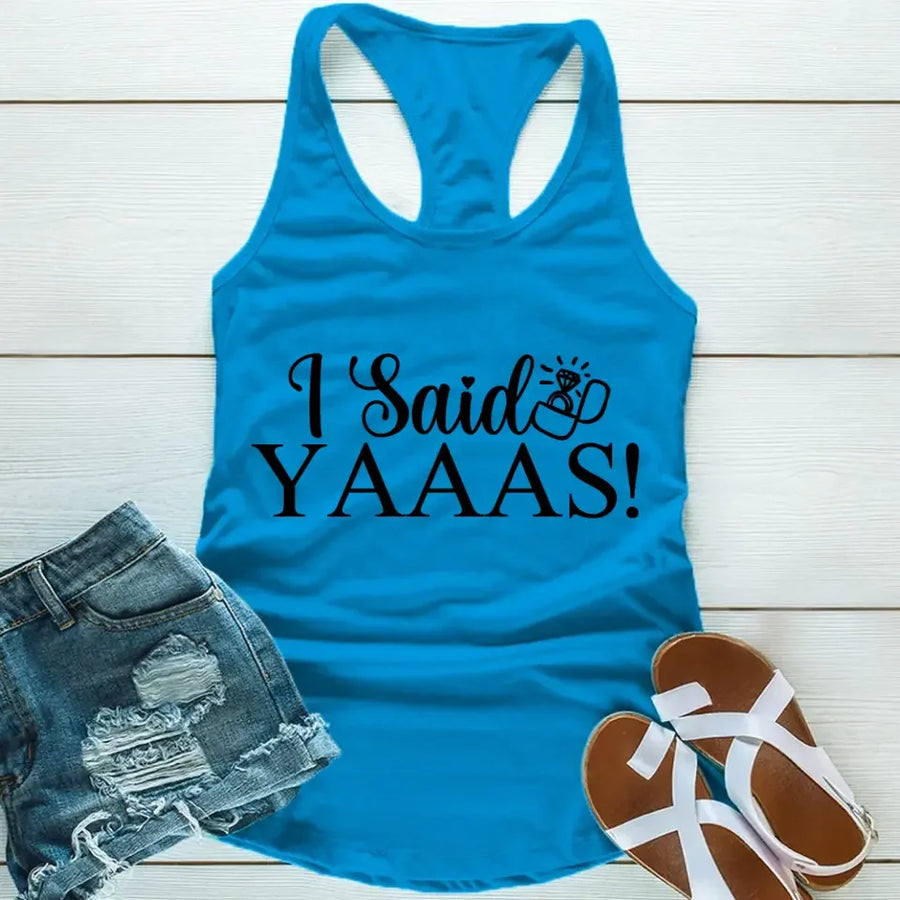 A Blue tank top with the text 'I Said YAAAS!' printed in black, placed on a wooden surface with blue denim shorts and white sandals beside it.