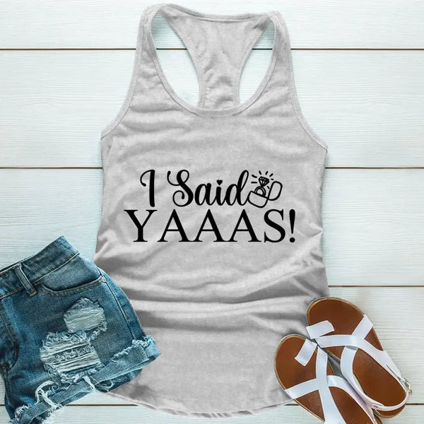 A grey tank top with the text 'I Said YAAAS!' printed in black, placed on a wooden surface with blue denim shorts and white sandals beside it.