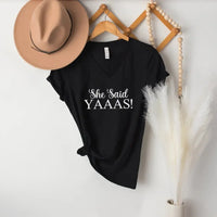 Black t-shirt with 'She Said YAAAS!' text, beige hat, and decorative elements on a white wall.