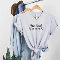 Gray t-shirt with 'She Said YAAAS!' text on a hanger with a denim jacket and hat in the background.