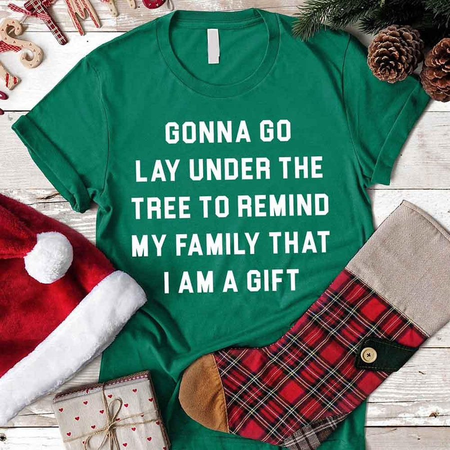 Green t-shirt with Lay Under A Tree Christmas text on a wooden surface with holiday items.