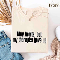 May include a person wearing an ivory colored sarcastic graphic t-shirt with the phrase “Muy Bonita But My Therapist Gave Up”.

