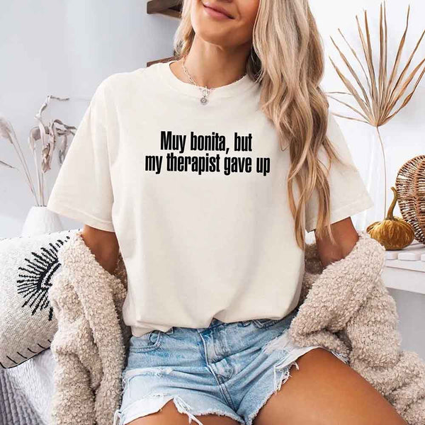 May include a person wearing an ivory colored sarcastic graphic t-shirt with the phrase “Muy Bonita But My Therapist Gave Up”.
