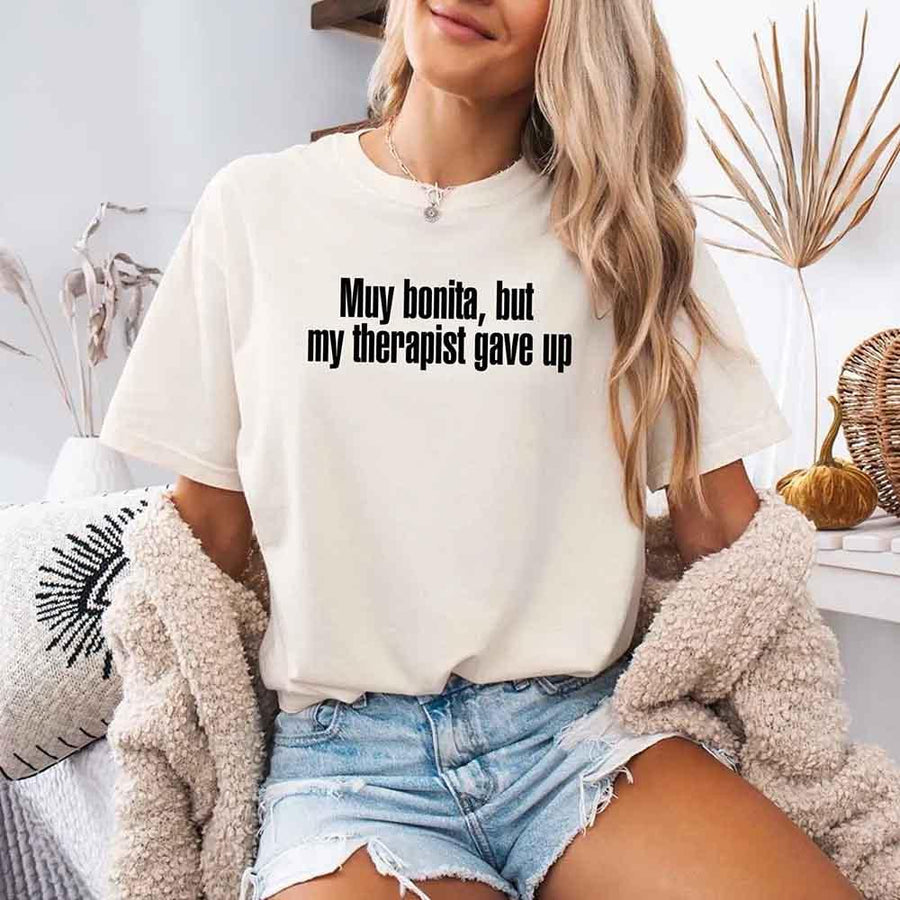 May include a person wearing an ivory colored sarcastic graphic t-shirt with the phrase “Muy Bonita But My Therapist Gave Up”.
