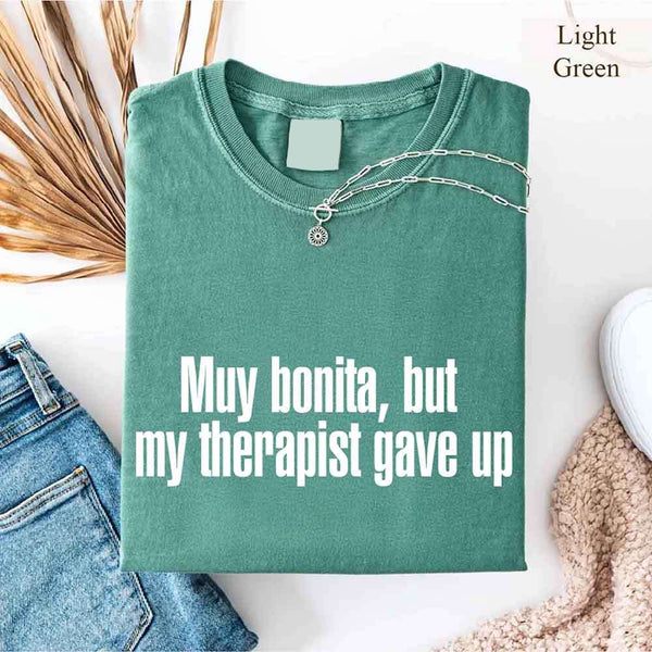 May include a person wearing a light green colored sarcastic graphic t-shirt with the phrase “Muy Bonita But My Therapist Gave Up”.
