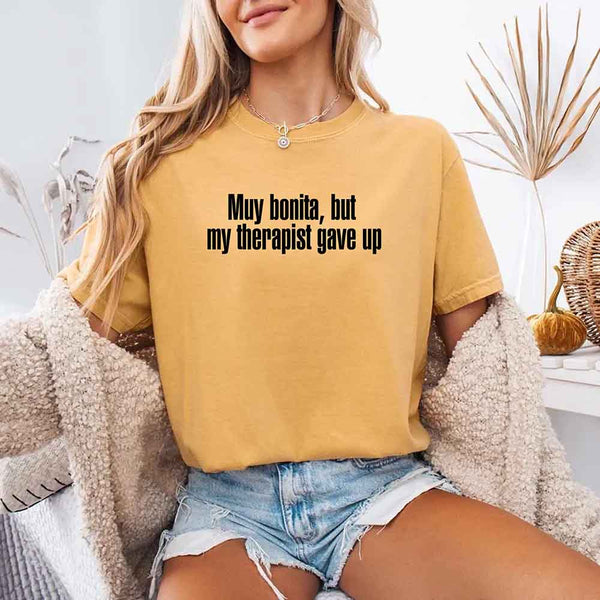 May include a person wearing a mustard colored sarcastic graphic t-shirt with the phrase “Muy Bonita But My Therapist Gave Up”.
