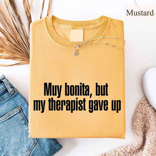 May include a person wearing a mustard colored sarcastic graphic t-shirt with the phrase “Muy Bonita But My Therapist Gave Up”.
