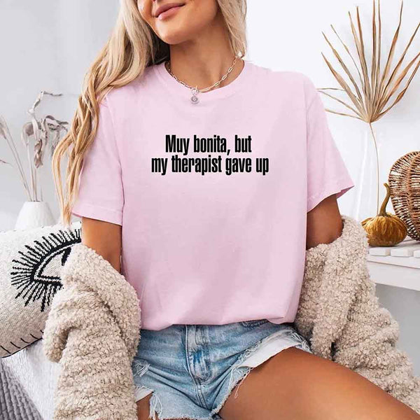 May include a person wearing a blossom colored sarcastic graphic t-shirt with the phrase “Muy Bonita But My Therapist Gave Up”.
