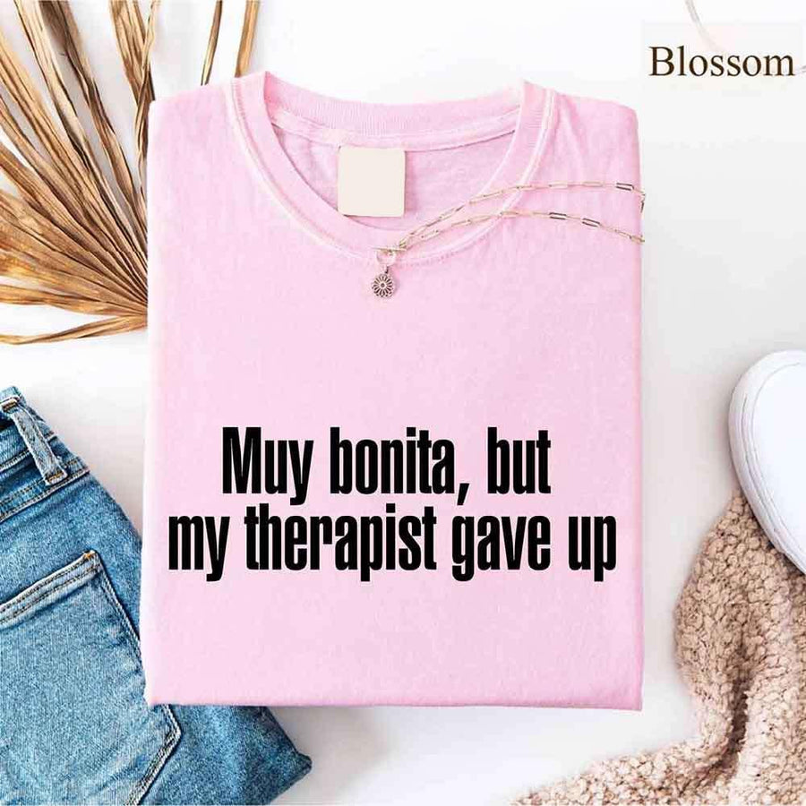 May include a person wearing a blossom colored sarcastic graphic t-shirt with the phrase “Muy Bonita But My Therapist Gave Up”.
