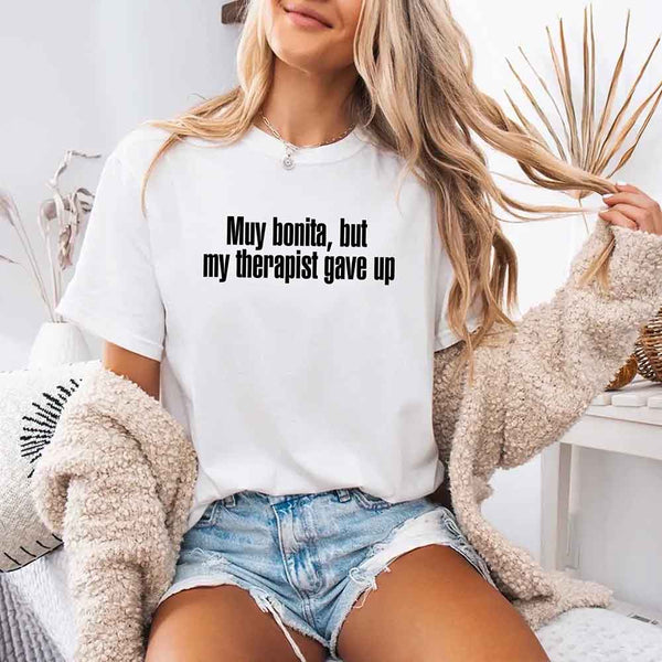 May include a person wearing a white colored sarcastic graphic t-shirt with the phrase “Muy Bonita But My Therapist Gave Up”.
