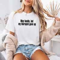 May include a person wearing a white colored sarcastic graphic t-shirt with the phrase “Muy Bonita But My Therapist Gave Up”.
