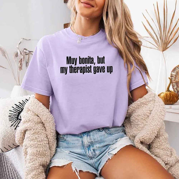 May include a person wearing an orchid colored sarcastic graphic t-shirt with the phrase “Muy Bonita But My Therapist Gave Up”.
