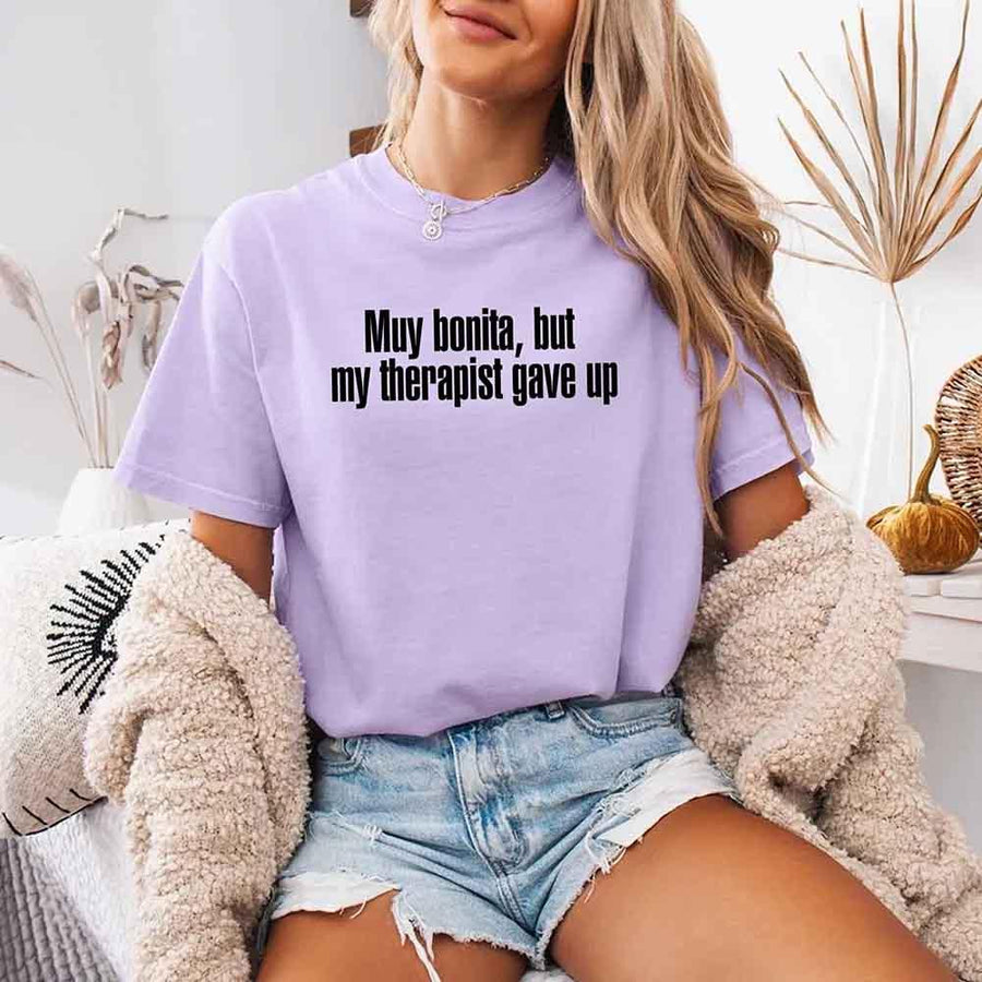 May include a person wearing an orchid colored sarcastic graphic t-shirt with the phrase “Muy Bonita But My Therapist Gave Up”.
