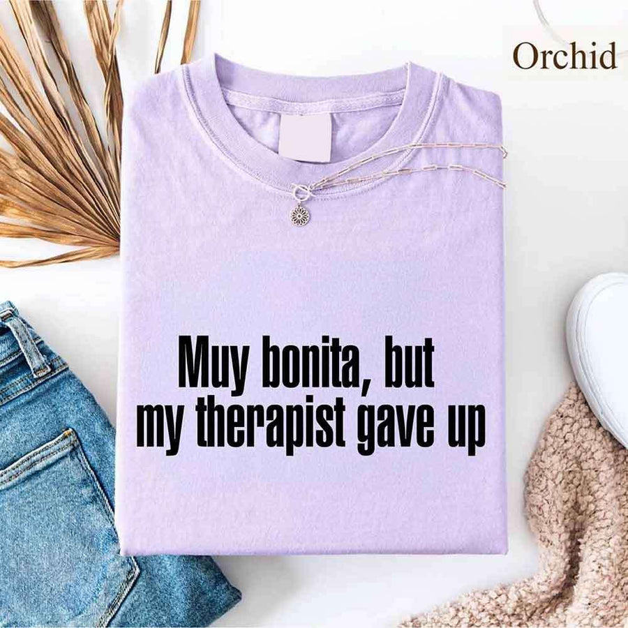 May include a person wearing an orchid colored sarcastic graphic t-shirt with the phrase “Muy Bonita But My Therapist Gave Up”.
