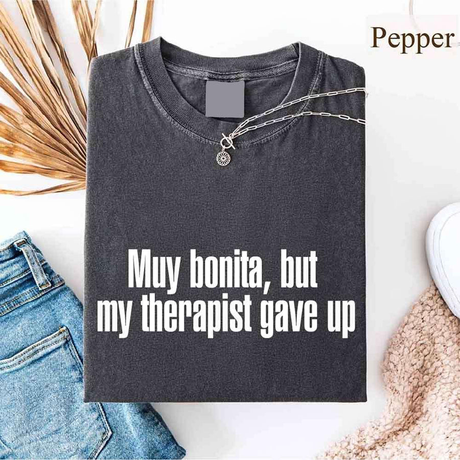 May include a person wearing a pepper colored sarcastic graphic t-shirt with the phrase “Muy Bonita But My Therapist Gave Up”.

