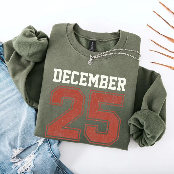 May include a retro-style Christmas sweatshirt featuring bold varsity lettering that reads “December 25” in green and red. The design has a vintage, slightly distressed look, giving it a nostalgic holiday feel perfect for festive winter wear.