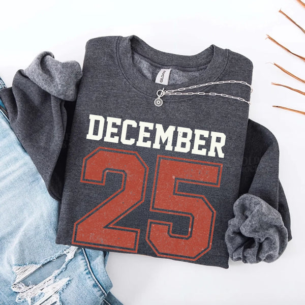 May include a retro-style Christmas sweatshirt featuring bold varsity lettering that reads “December 25” in green and red. The design has a vintage, slightly distressed look, giving it a nostalgic holiday feel perfect for festive winter wear.