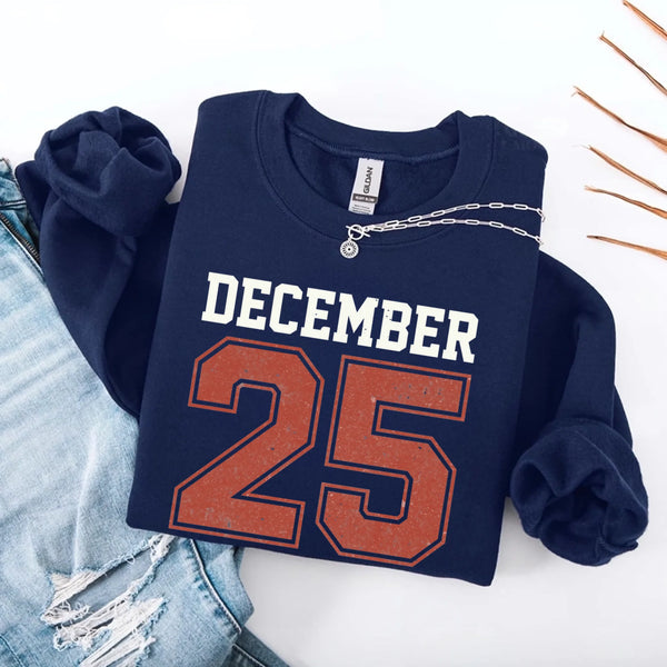 May include a retro-style Christmas sweatshirt featuring bold varsity lettering that reads “December 25” in green and red. The design has a vintage, slightly distressed look, giving it a nostalgic holiday feel perfect for festive winter wear.