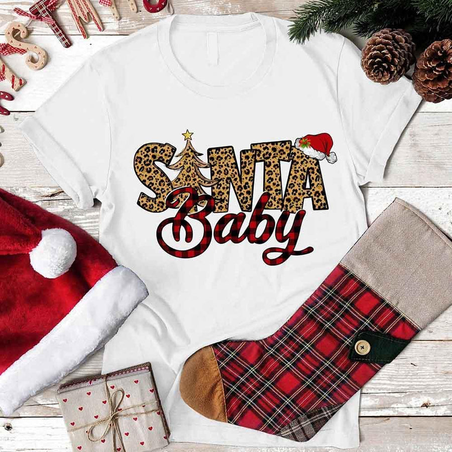 White t-shirt with 'Santa Baby' design, red Santa hat, plaid stocking, and pine cones on a wooden surface.