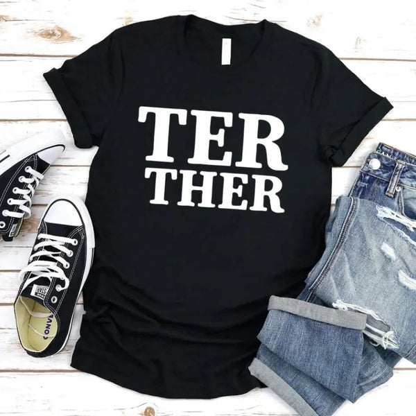 Black t-shirt with 'TER THER' text, paired with black sneakers and denim shorts on a wooden surface.