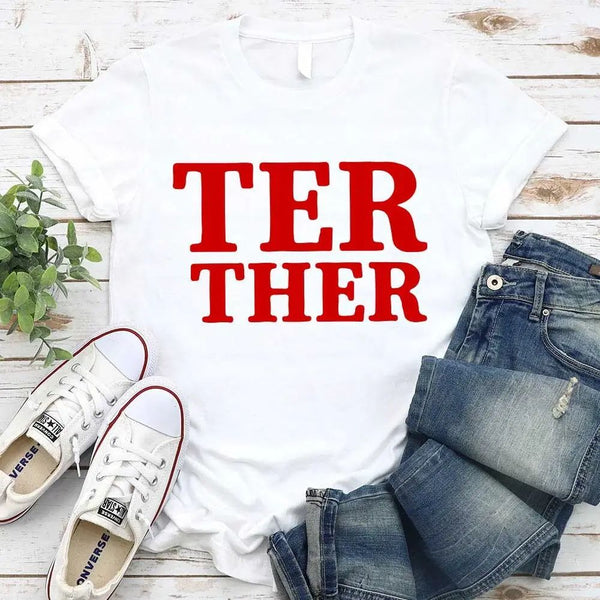 White t-shirt with red text, blue jeans, white sneakers, and a plant on a wooden surface.