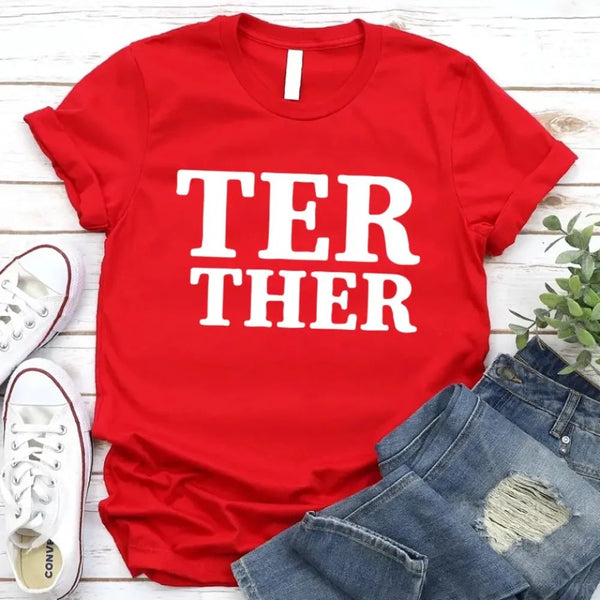 Red t-shirt with 'TER THER' text, white sneakers, and jeans on a wooden surface.