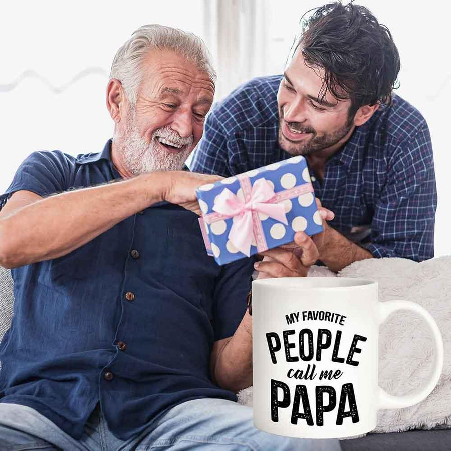 Two men sitting on a couch, one giving a gift to the other with a mug in front that says 'My favorite people call me Papa'.