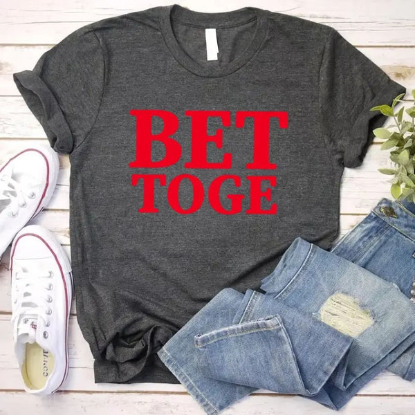 Gray t-shirt with red text 'BET TOGE', white sneakers, and blue jeans on a wooden surface.