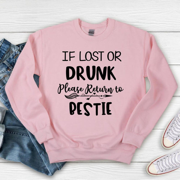 Pink sweatshirt with text 'If lost or drunk, please return to bestie' on a wooden surface with jeans and shoes.