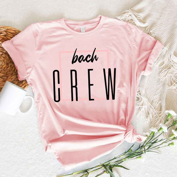 Pink t-shirt with 'bach CREW' text on a textured surface with flowers and a mug.