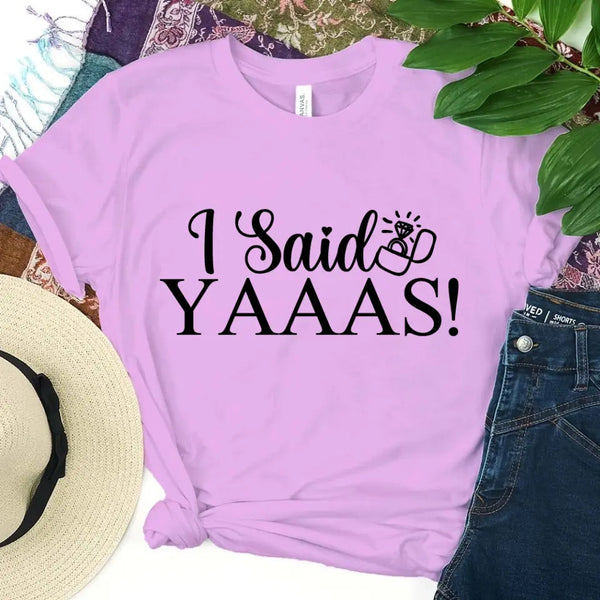 A lavender t-shirt with 'I Said YAAAS!' text, placed on a surface with a hat and jeans.