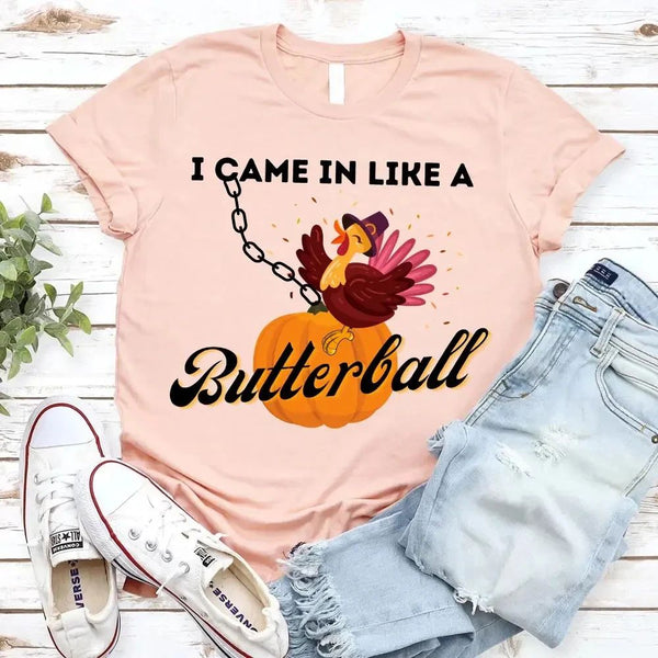 May include a casual crew-neck T-shirt with a playful Thanksgiving graphic showing a turkey stepping onto or perched on a pumpkin, surrounded by leaves or autumn motifs, with bold front text reading “I Came In Like A Butterball.”