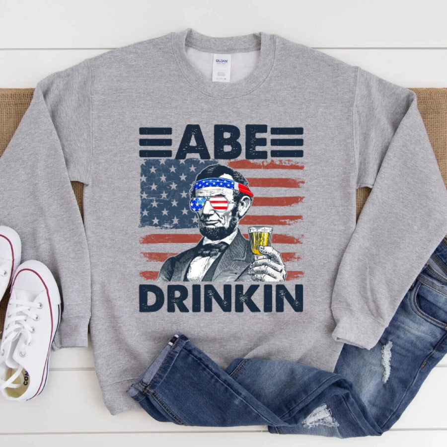 Gray sweatshirt with a graphic of a historical figure holding a drink, American flag, and text 'ABE DRINKIN'.