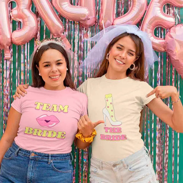 Two women wearing 'Team Bride' and 'The Bride' shirts with pink balloons and streamers in the background.