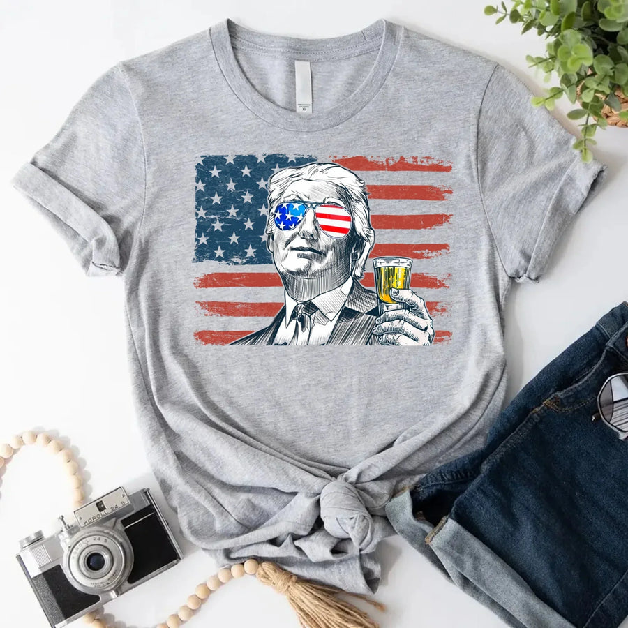 Gray t-shirt with a graphic of a person holding a drink and an American flag, placed on a white surface with accessories.