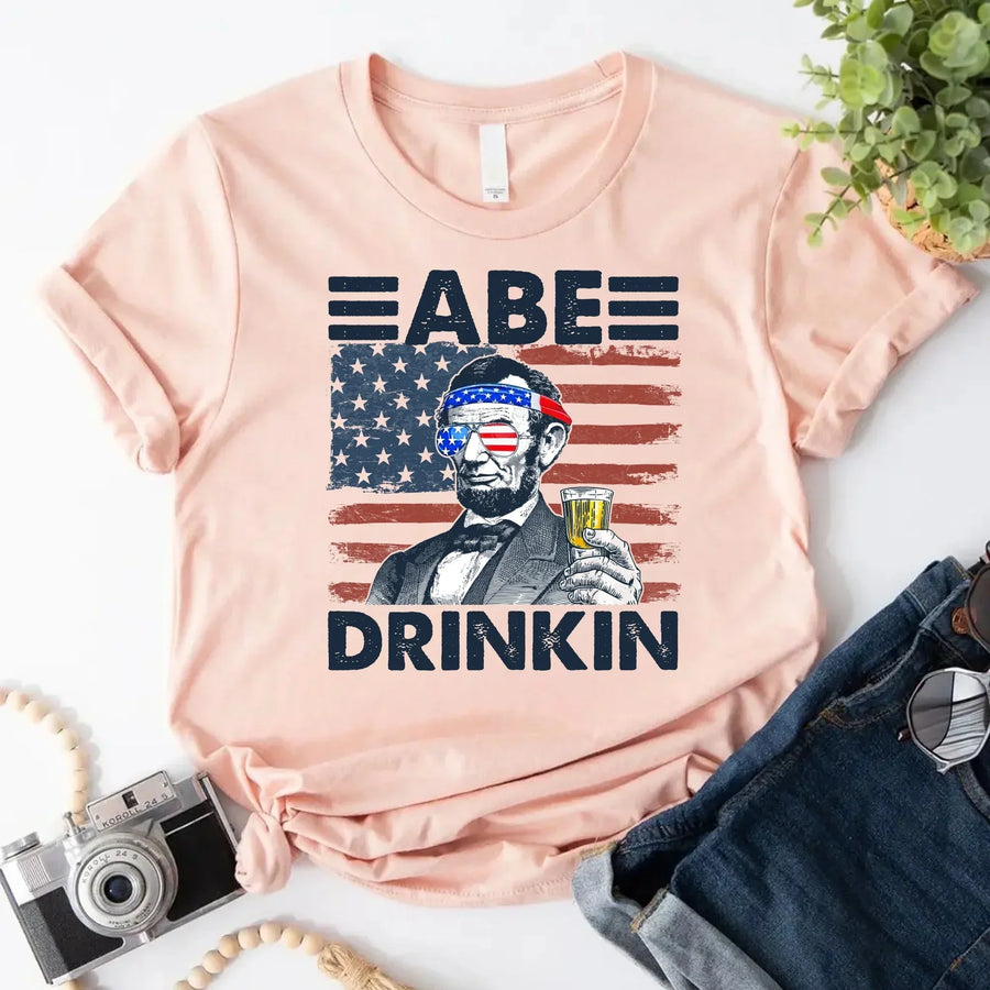 Peach custom tee with Abraham Lincoln, American flag, sunglasses, drink, patriotic graphic.
