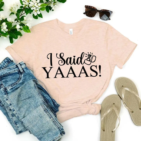 Beige t-shirt with 'I Said YAAAS!' text, paired with denim shorts and sandals on a white background.