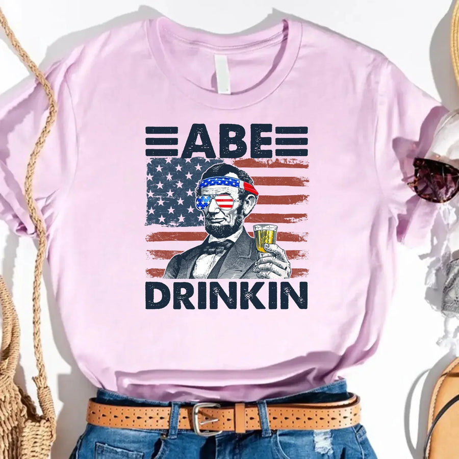 Pink graphic tee with Abe Lincoln holding a drink, American flag sunglasses, patriotic design