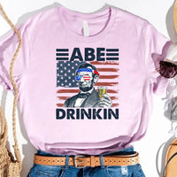 Pink graphic tee with Abe Lincoln holding a drink, American flag sunglasses, patriotic design