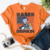 Orange custom graphic tee with Abraham Lincoln, sunglasses with American flag, and drink, styled with denim shorts.
