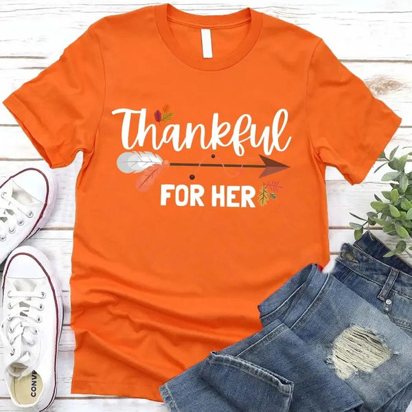 May include two matching Thanksgiving-themed T-shirts laid flat, one black reading “Thankful For Her” and one orange reading “Thankful For Him.” Both designs feature autumn leaves, feathers, and an arrow motif, surrounded by small pumpkins and fall decor.
