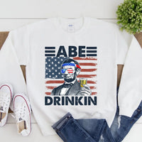 White sweatshirt with a graphic design featuring a person holding a drink, American flag, and text 'ABE DRINKIN'.