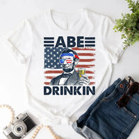 White custom patriotic tee with cartoon Abraham Lincoln holding a beer, USA flag background, styled with denim shorts.