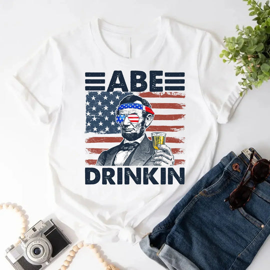 White custom patriotic tee with cartoon Abraham Lincoln holding a beer, USA flag background, styled with denim shorts.