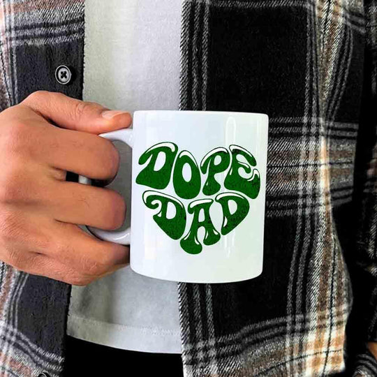 Person holding a white mug with 'DOPE DAD' in green text, wearing a plaid shirt.