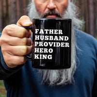 Person holding a black mug with text 'FATHER HUSBAND PROVIDER HERO KING' against a blurred background.