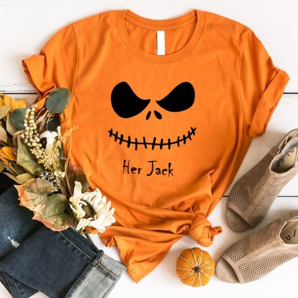 Orange t-shirt with pumpkin face design and 'Her Jack' text, surrounded by autumn-themed items on a white surface.