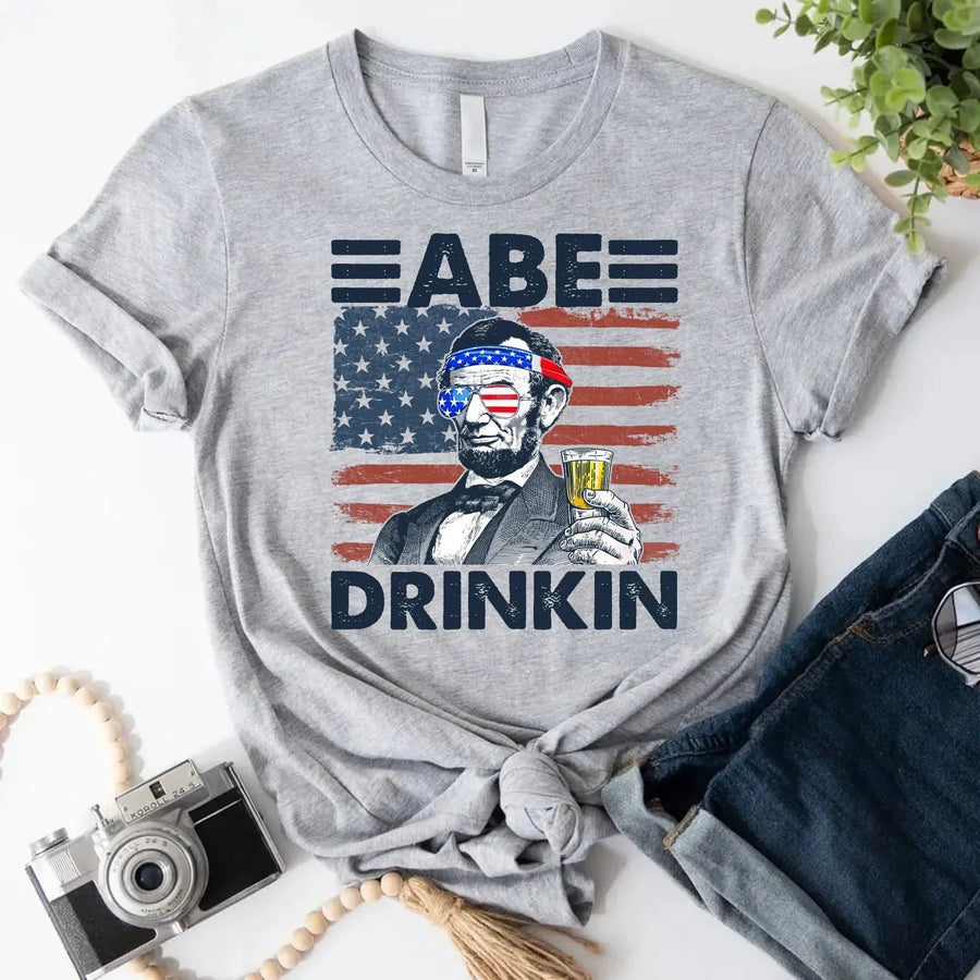 Gray graphic t-shirt with Abraham Lincoln holding a drink, American flag background, patriotic design