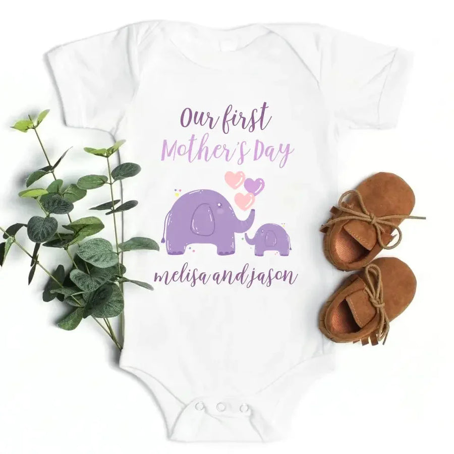 Baby onesie with 'Our first Mother's Day' text and elephant design, paired with baby shoes and greenery on a white background.