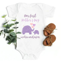 Baby onesie with 'Our first Mother's Day' text and elephant design, paired with baby shoes and greenery on a white background.